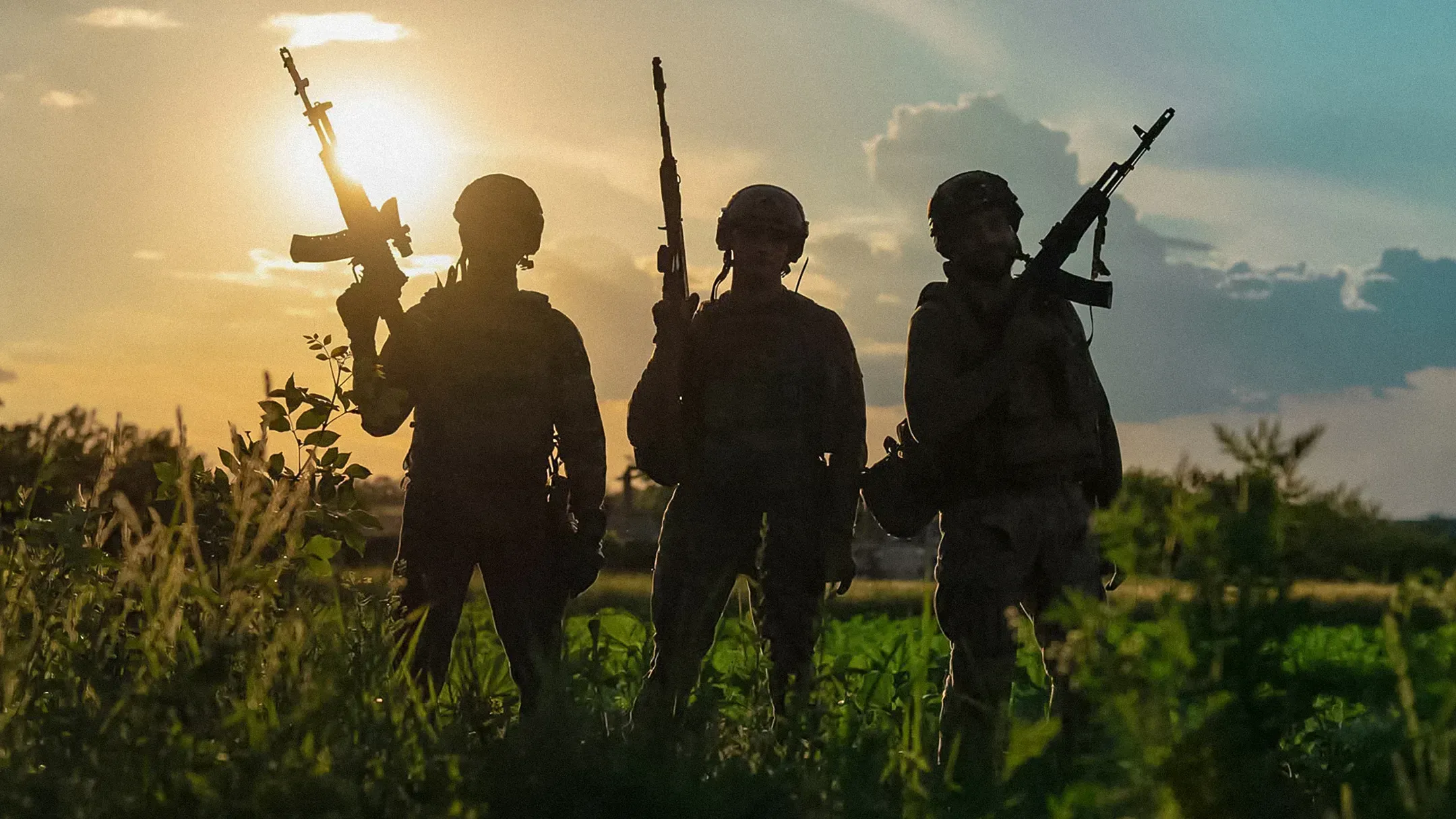 Three service members in military gear holding weapons stand in a field against an evening or morning sky
