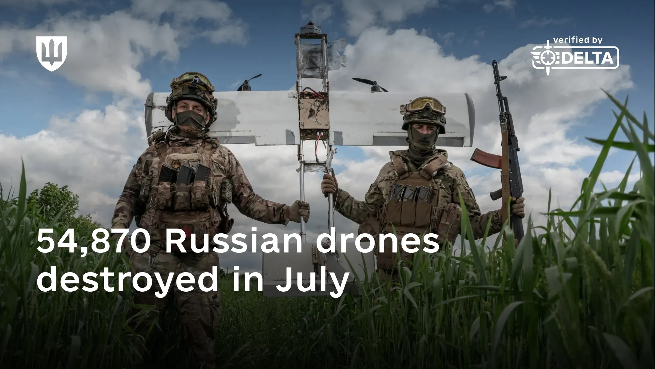 Two service members in camouflage and helmets hold a damaged fixed-wing drone upright between them in tall grass, one of them carrying a rifle