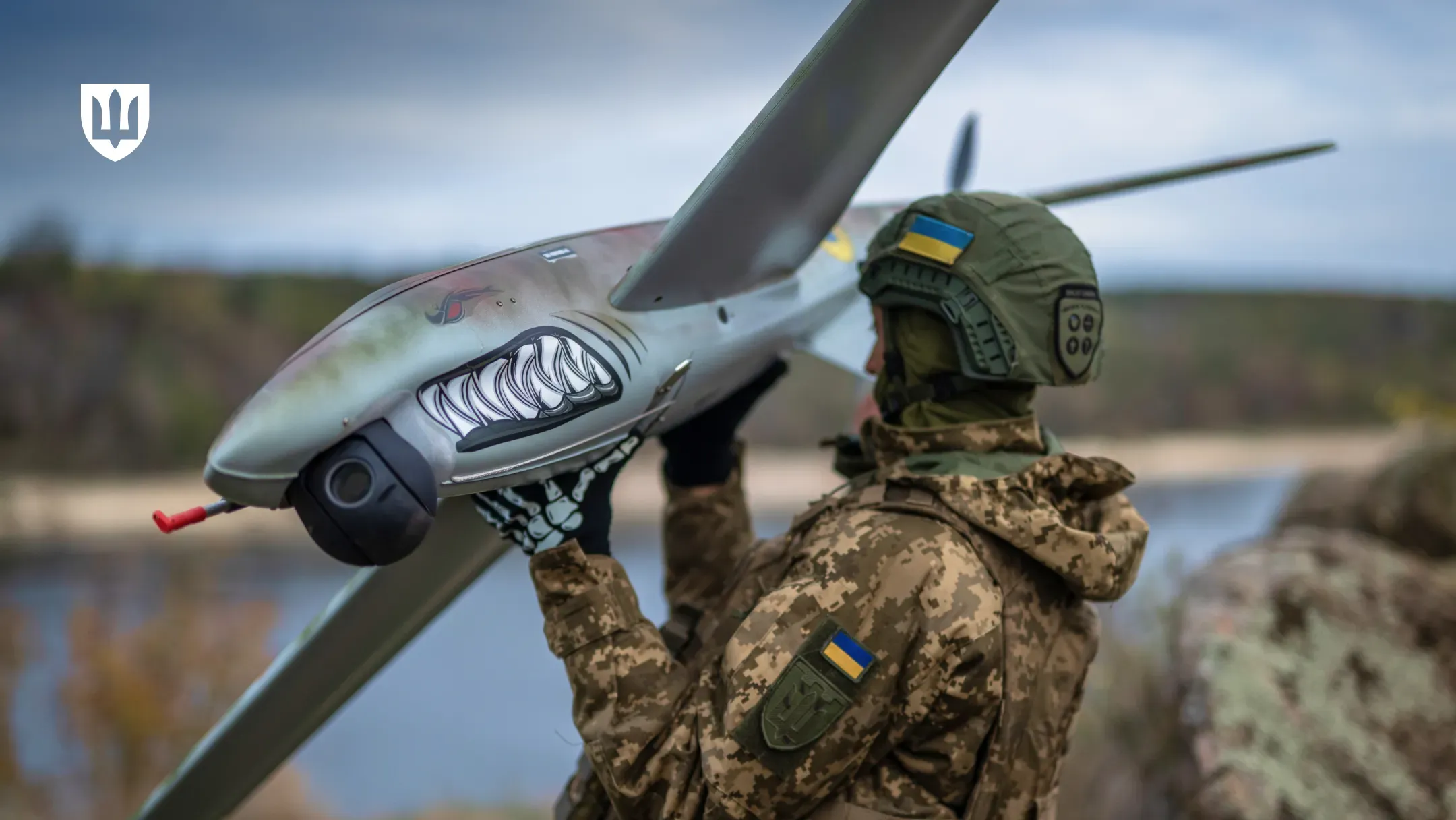 Український військовослужбовець-оператор БпАК у камуфляжі та шоломі тримає в руках вітчизняний безпілотник літакового типу SHARK, готуючи його до польоту. На носовій частині сірого дрона нанесений впізнаваний малюнок — хижа паща акули