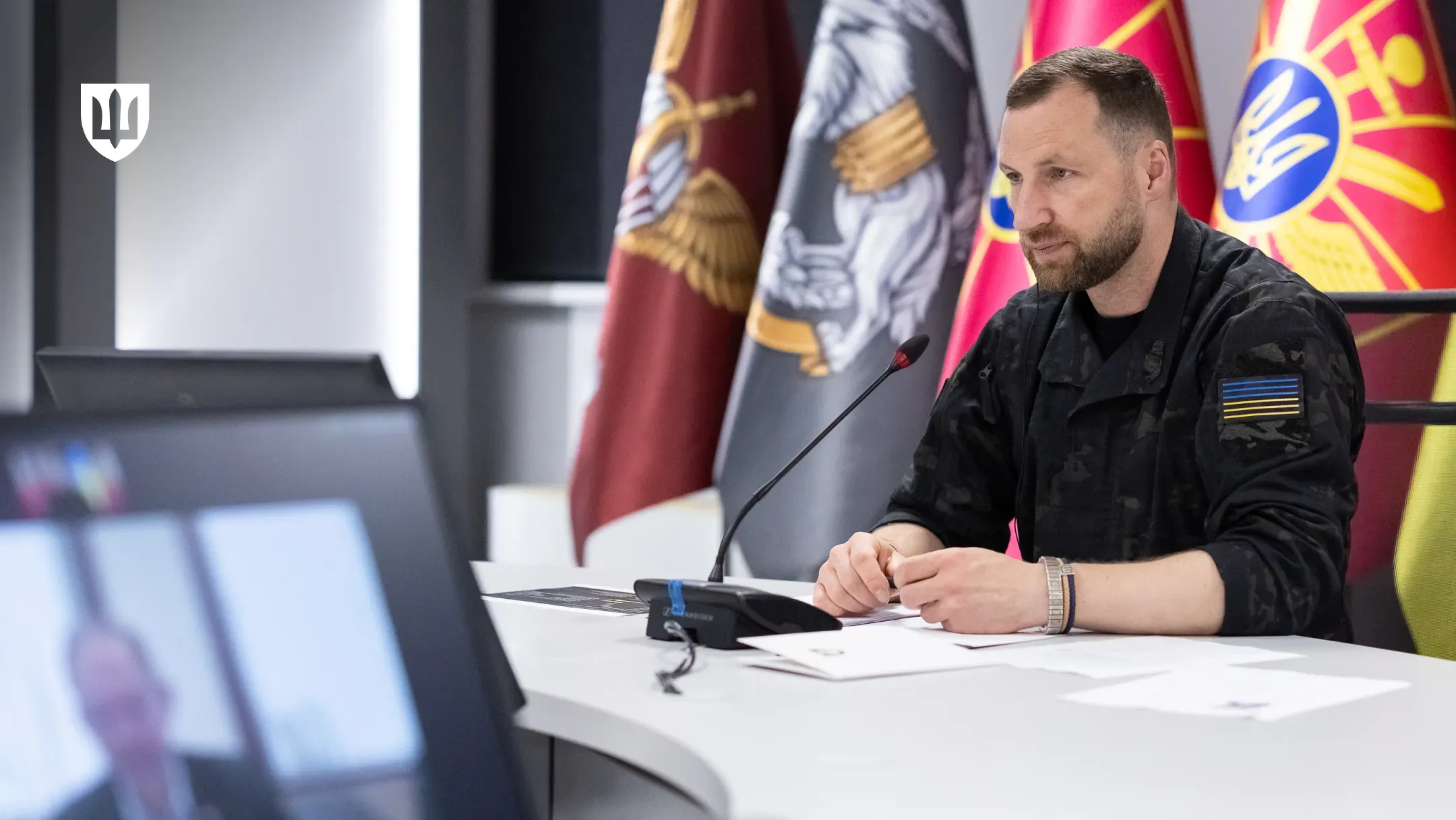 Міністр оборони України Євгеній Хмара сидить за столом у залі засідань під час телефонної розмови у форматі відеозв’язку. Перед ним ноутбук, мікрофон і документи, позаду розміщені прапори та символіка Міністерства оборони України.