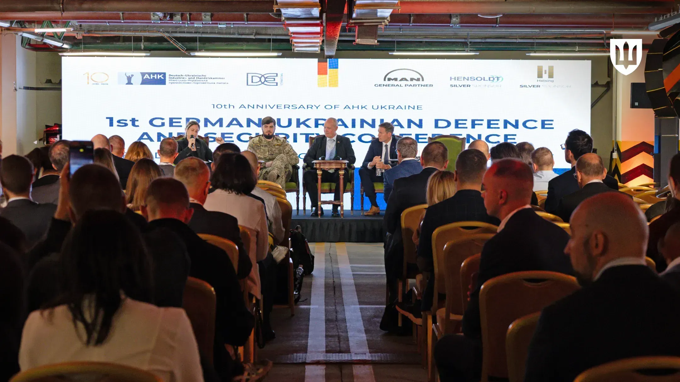 Panel discussion on stage under a banner reading “10th Anniversary of AHK Ukraine, 1st German-Ukrainian Defence and Security Conference”, with the audience in the foreground