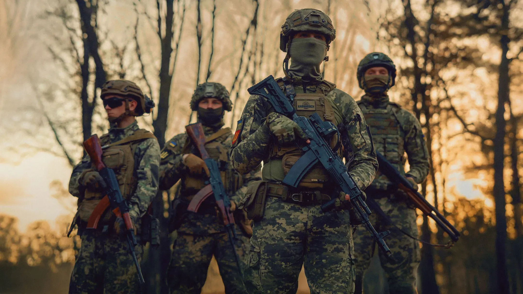 Four Ukrainian soldiers in full gear stand in a forest at a position against the backdrop of a sunset