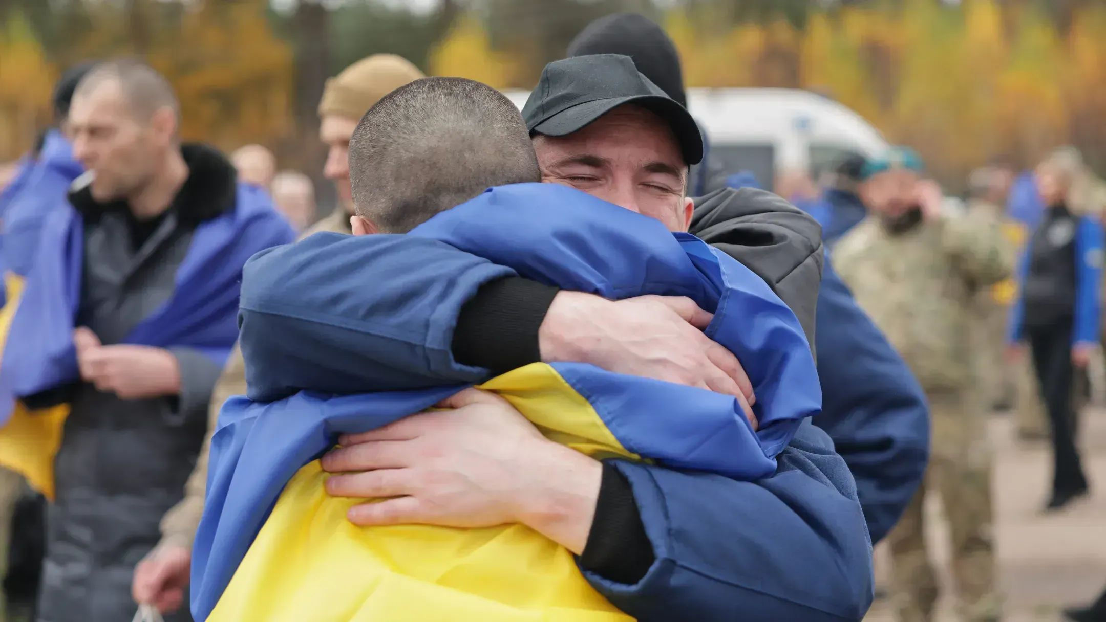 A Ukrainian freed from captivity, wrapped in a Ukrainian flag, embraces a man in a cap, with other freed Ukrainians wrapped in Ukrainian flags behind them