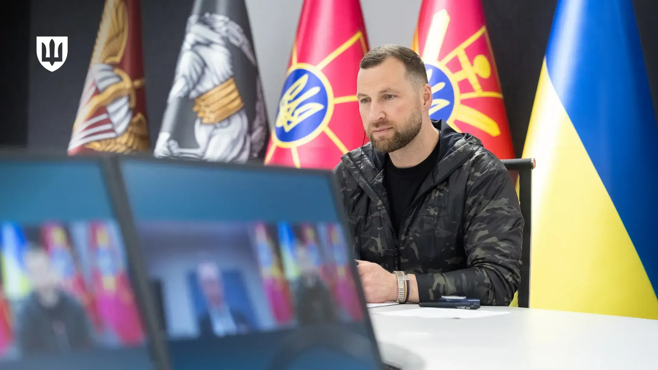 Evgeniy Khmara at a table during a video call with Tore Sandvik, with the Ukrainian flag and military flags behind him and monitors in the foreground